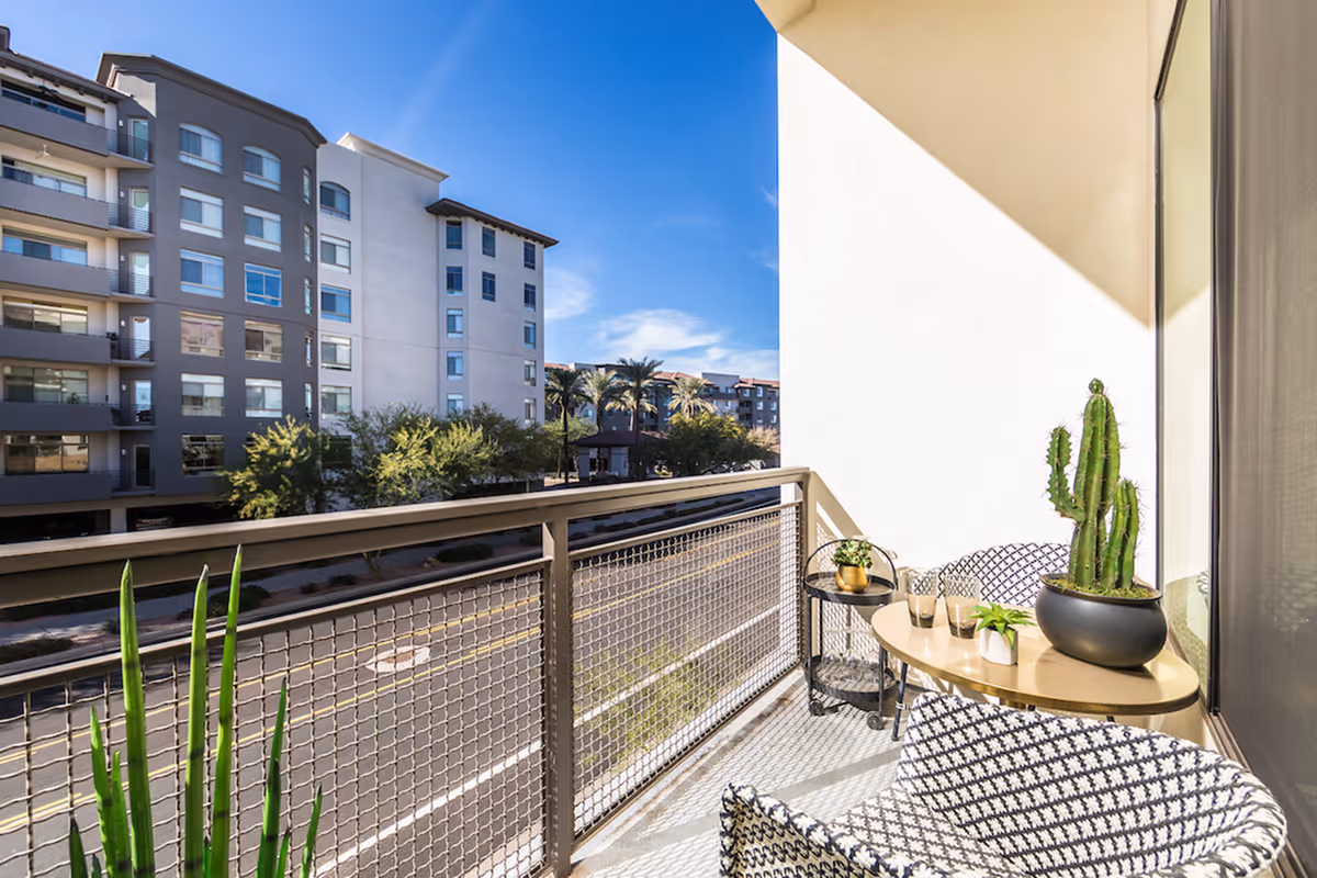 A sunny balcony with a patterned chair, a small round table holding a potted cactus and two glasses, and a small side table with another plant. The balcony overlooks a street with trees and a multi-story residential building under a clear blue sky.