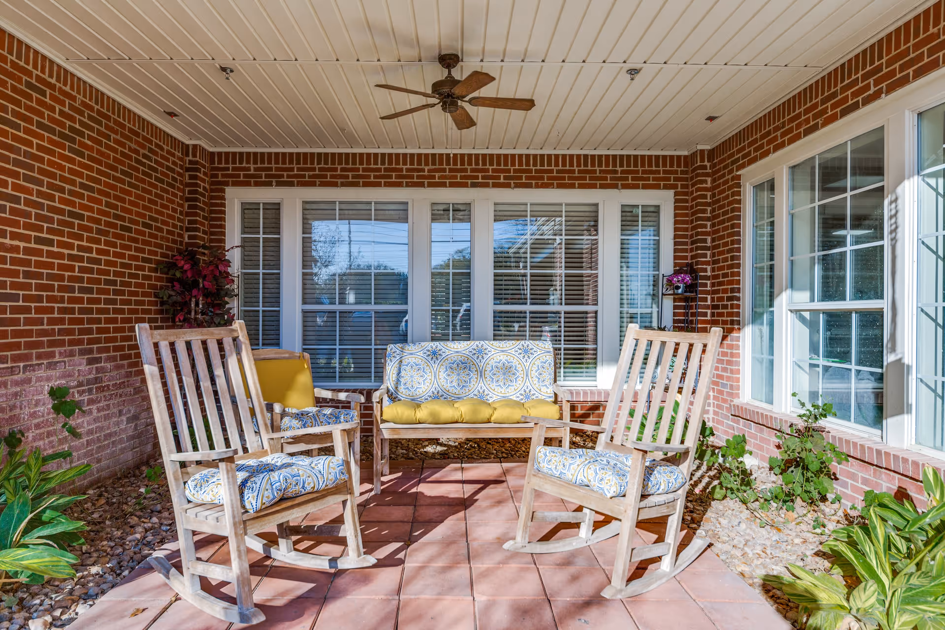 A covered outdoor patio area with two wooden rocking chairs and a cushioned bench with patterned pillows. The patio has a brick wall, large windows with white frames, a ceiling fan, and some potted plants along the edges.