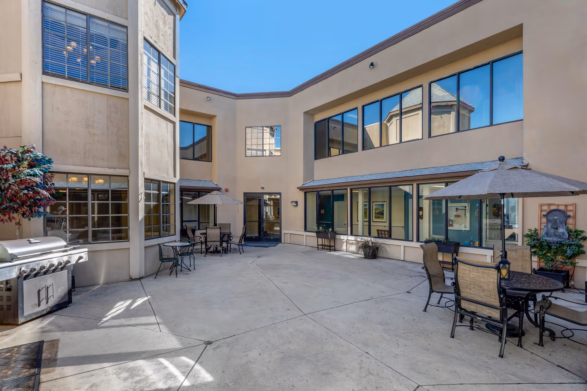 Sunlit central courtyard with patio tables and umbrellas, a grill, and two‑story windows surrounding the space.