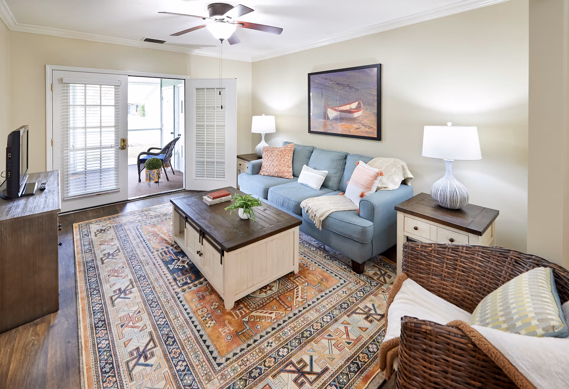 A cozy living room with a blue sofa adorned with pillows and a throw blanket, a wooden coffee table with a small plant and books, a wicker chair with cushions, two side tables each with a lamp, a ceiling fan with light, and French doors leading to a small outdoor seating area with two chairs and a small table.
