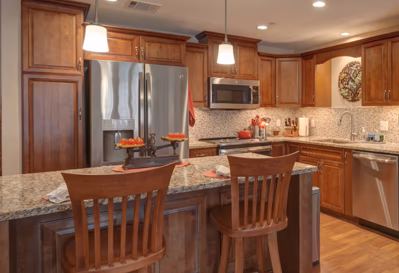 A modern kitchen with wooden cabinets, granite countertops, and stainless steel appliances including a refrigerator, microwave, oven, and dishwasher. Two wooden chairs are placed at the kitchen island, which has a decorative scale with tomatoes on it. The backsplash features small mosaic tiles, and two pendant lights hang above the island.