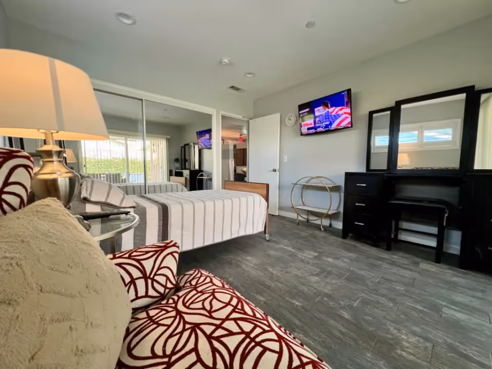 A bright bedroom with a single bed covered in a striped bedspread, a patterned armchair with a beige pillow, a bedside table with a lamp, a wall-mounted TV, a clock, a rolling cart, and a black dresser with a mirror and a stool. The room has a sliding glass door with vertical blinds and a gray tiled floor.