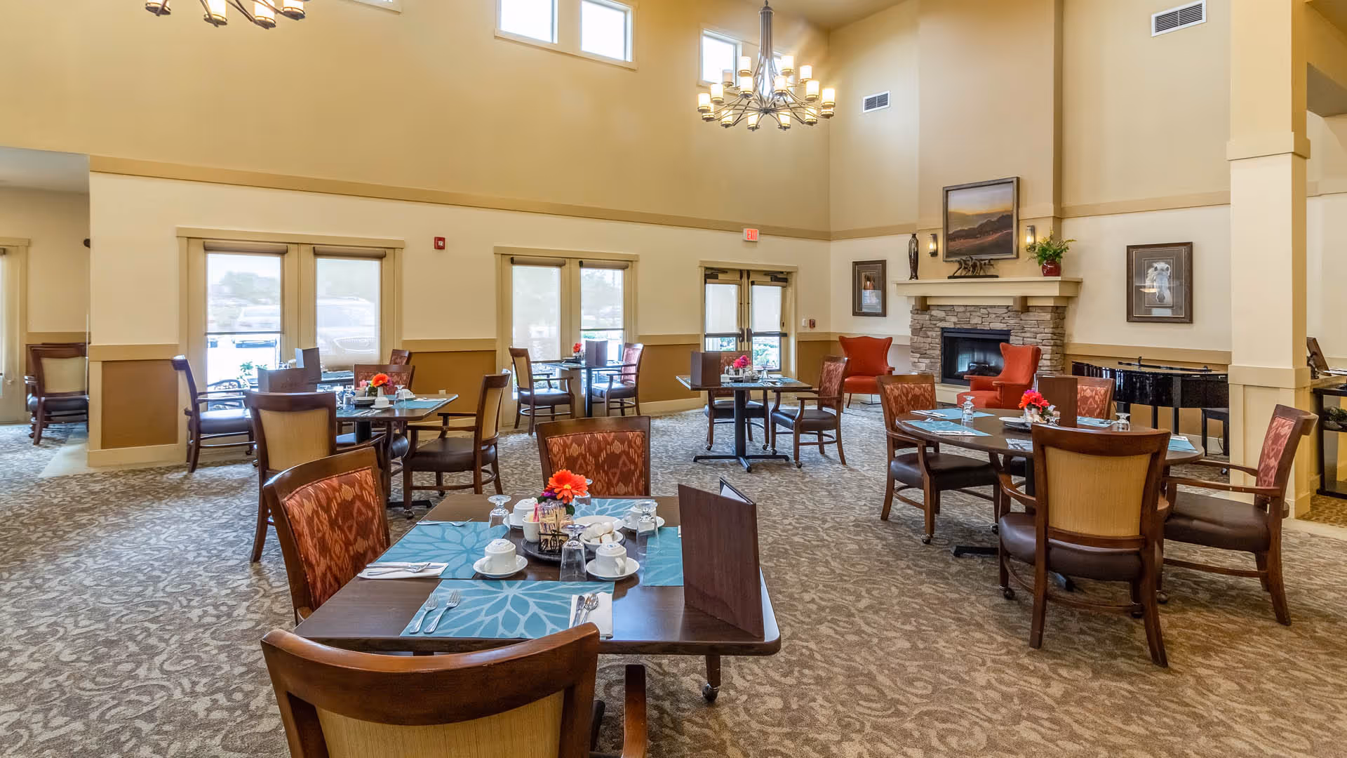 A spacious dining room with multiple tables set for meals, each with placemats, cups, and utensils. The room features high ceilings with a chandelier, large windows allowing natural light, a stone fireplace with artwork above it, and comfortable chairs around the tables. The decor is warm and inviting with beige walls and patterned carpet.