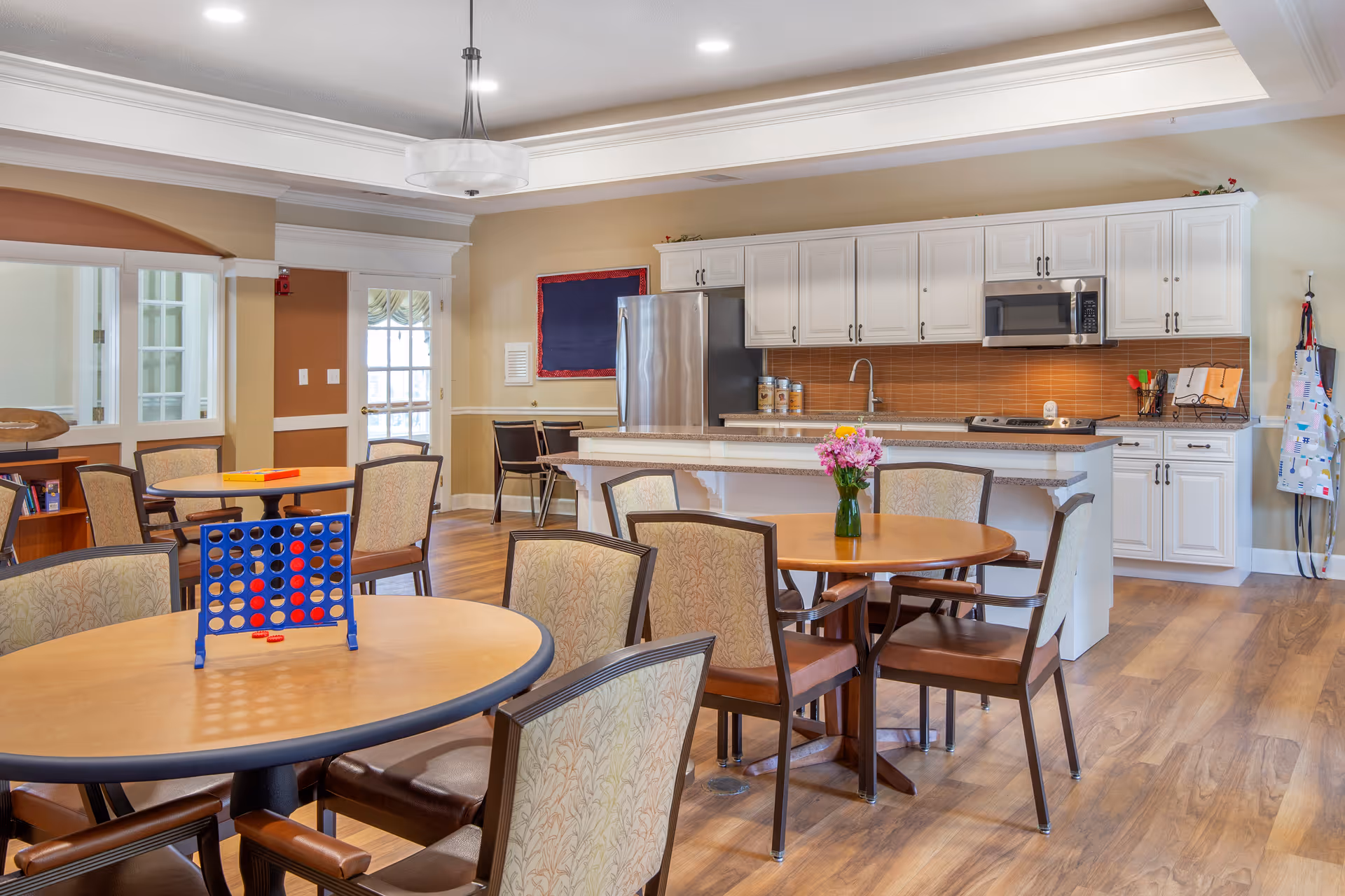 A bright and spacious common area featuring a kitchen with white cabinets, a stainless steel refrigerator, microwave, and stove. Several round tables with cushioned chairs are arranged around the room, one table has a Connect Four game set up, and another has a vase with pink flowers. The room has wood flooring and a ceiling light fixture.