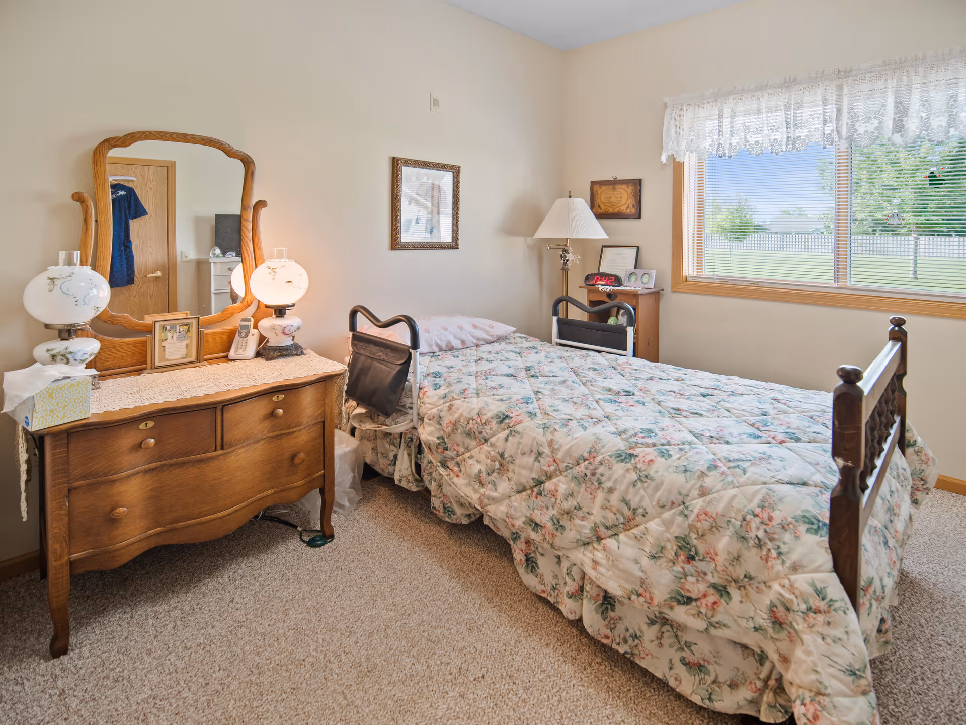 A cozy bedroom with a single bed covered in a floral quilt. The bed has safety rails on both sides. Next to the bed is a wooden dresser with a large mirror, two vintage-style lamps, a tissue box, and a cordless phone. A window with lace curtains lets in natural light and shows a green lawn and trees outside. The room has beige walls and carpeted floor.