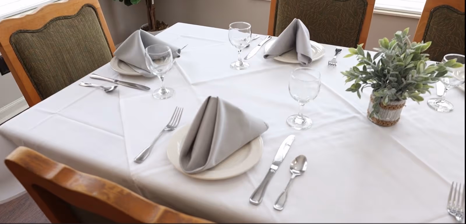 A dining table set for four with white tablecloth, neatly folded gray napkins on white plates, clear water glasses, silverware, and a small potted green plant centerpiece. Wooden chairs with green cushioned backs surround the table.