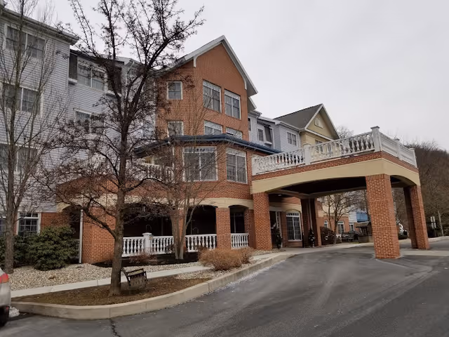 Exterior view of a multi-story senior living facility building with a covered entrance supported by brick columns. The building features a mix of brick and siding with multiple windows and a small landscaped area with trees and shrubs in front.