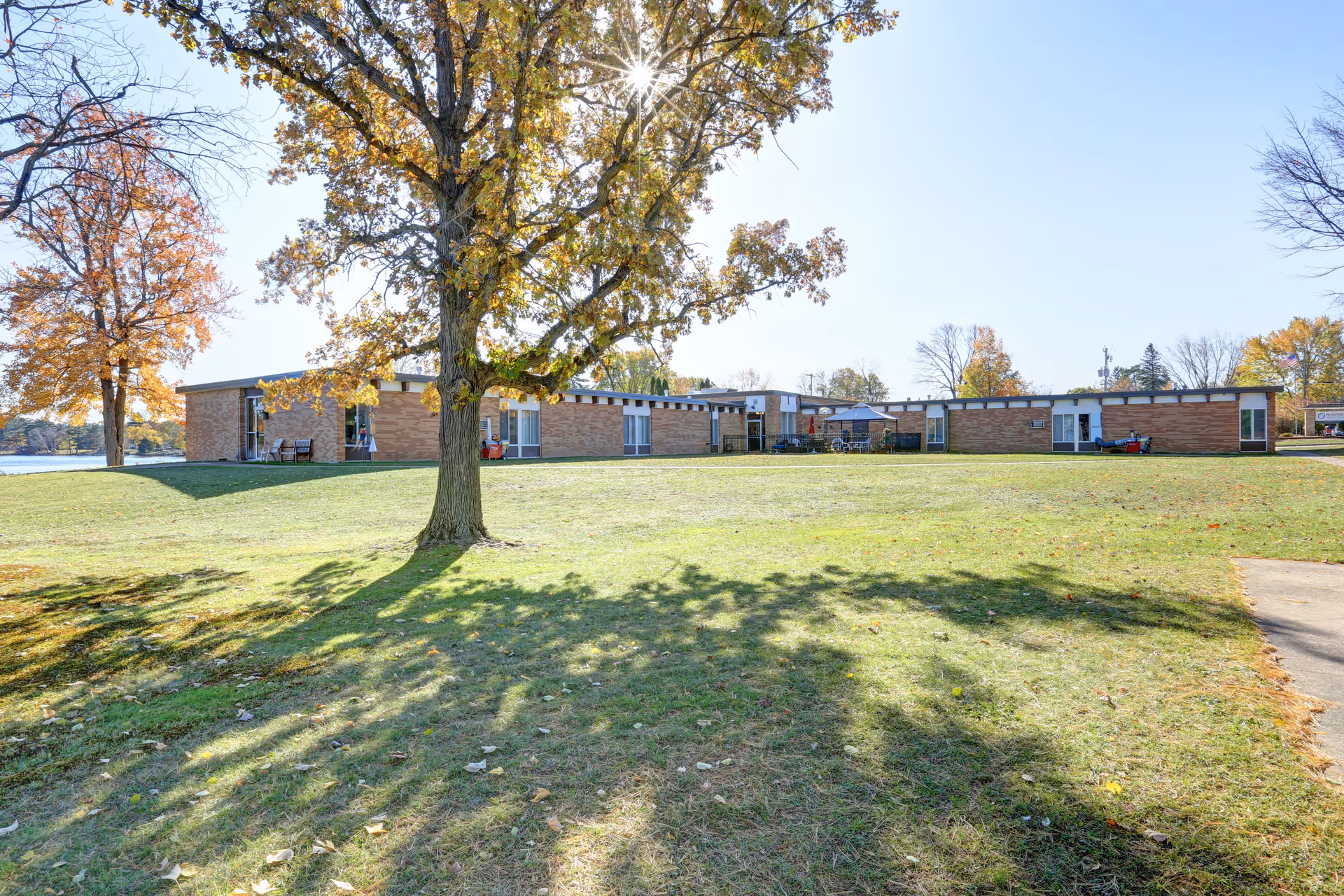 Single-story brick senior living facility building seen from a grassy lawn with a large tree in the foreground and water at the left.