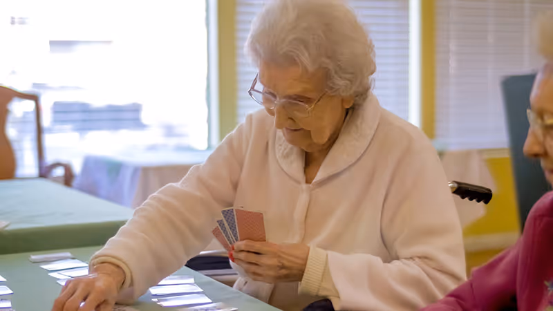 An elderly woman with white hair and glasses is sitting at a table playing a card game. She is wearing a light-colored sweater and holding several cards in her hand while looking at the cards on the table. Another elderly person is partially visible sitting next to her.