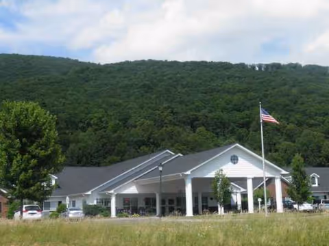Front view of Forest Ridge Assisted Living building with entrance, flag, cars, and wooded hillside.