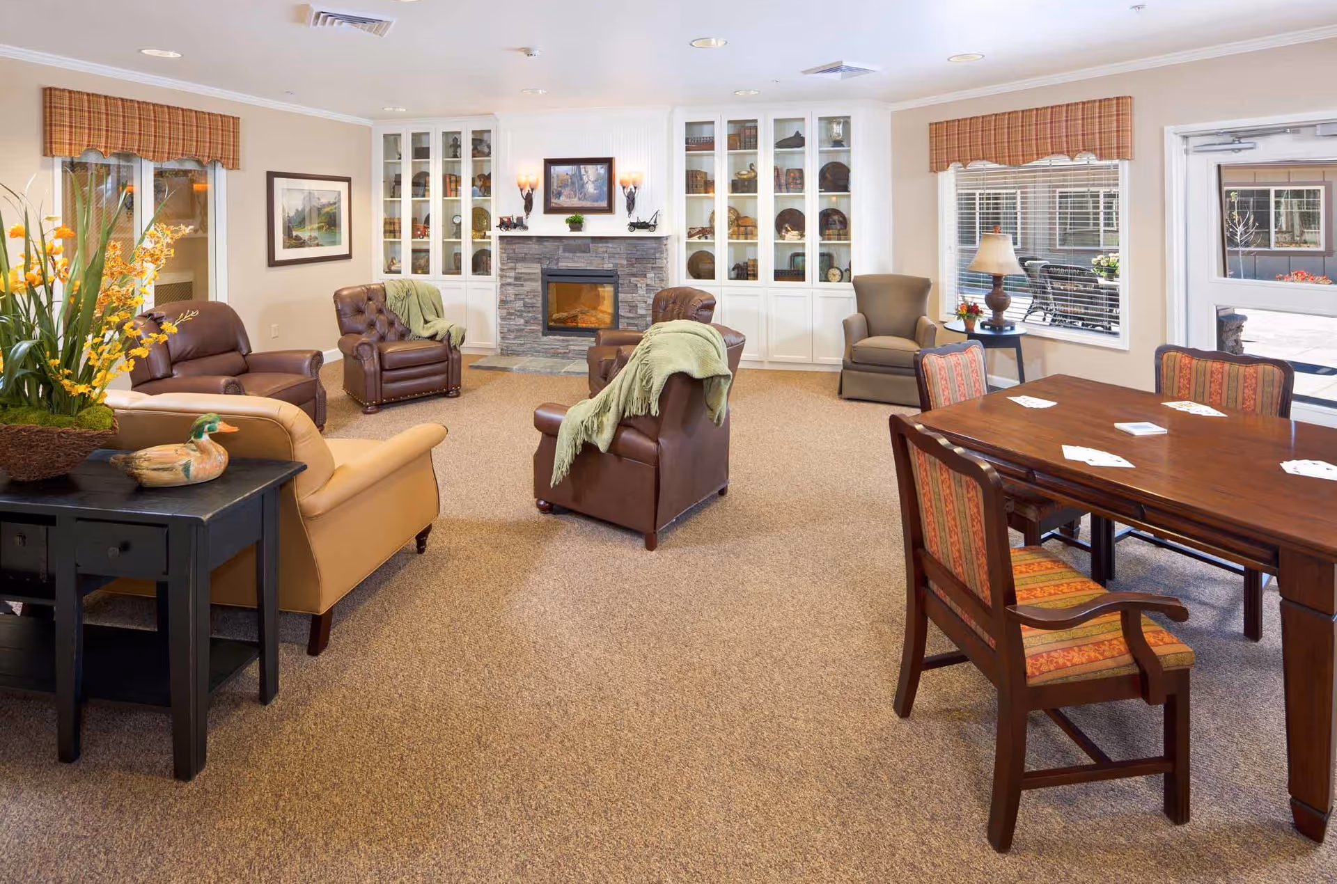 Bright communal living room with armchairs arranged around a fireplace and a dining table at the right.