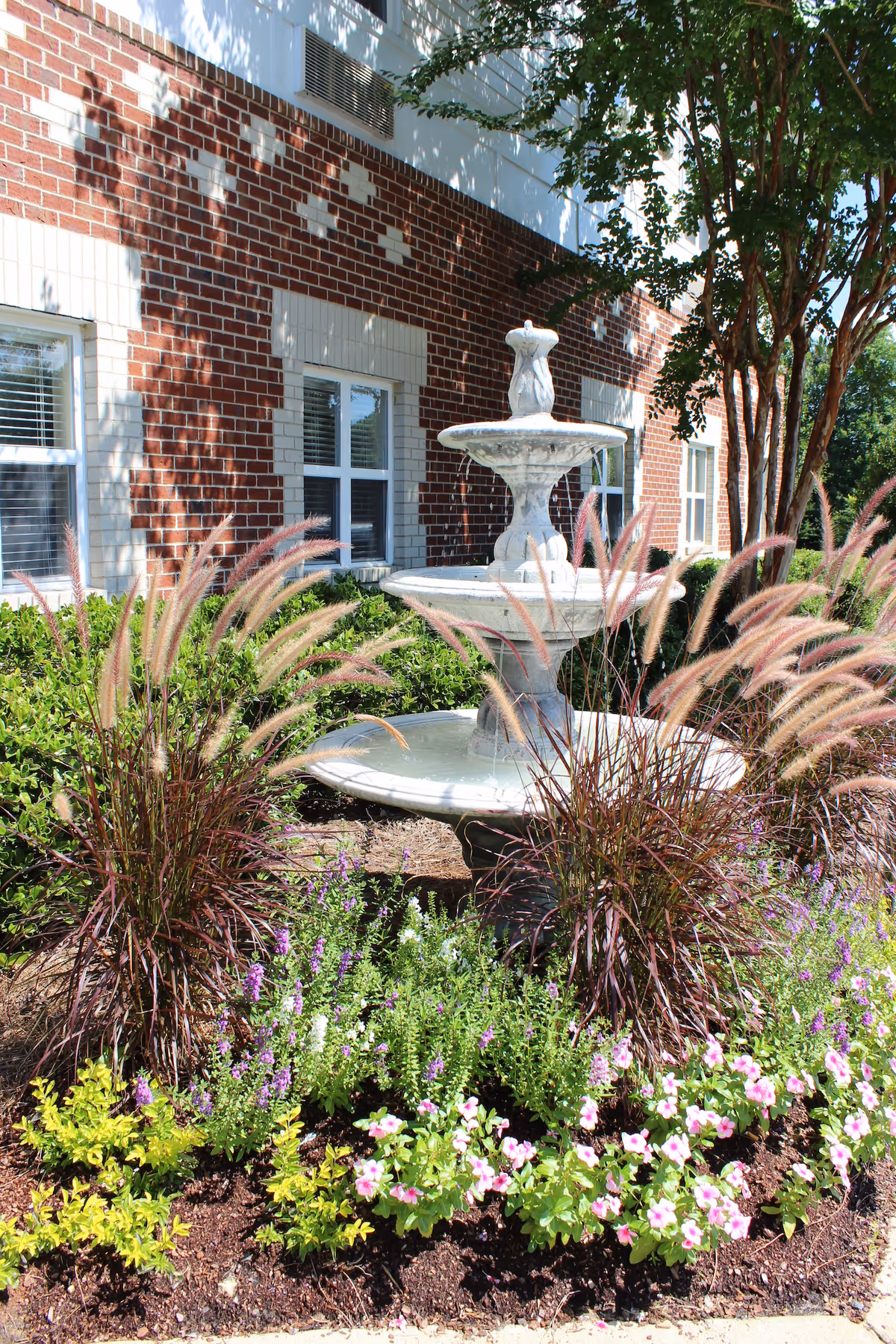 Outdoor garden area at Morningview at Irving Park featuring a white tiered water fountain surrounded by ornamental grasses, flowering plants, and shrubs in front of a red brick building with white-trimmed windows.