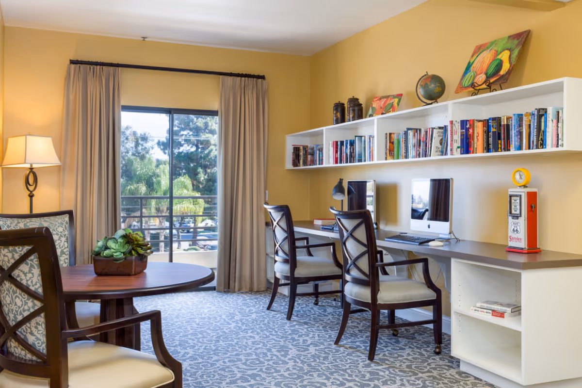Bright common area with a round table and chairs, a long desk with two computers beneath wall bookshelves, and a window with curtains overlooking trees.