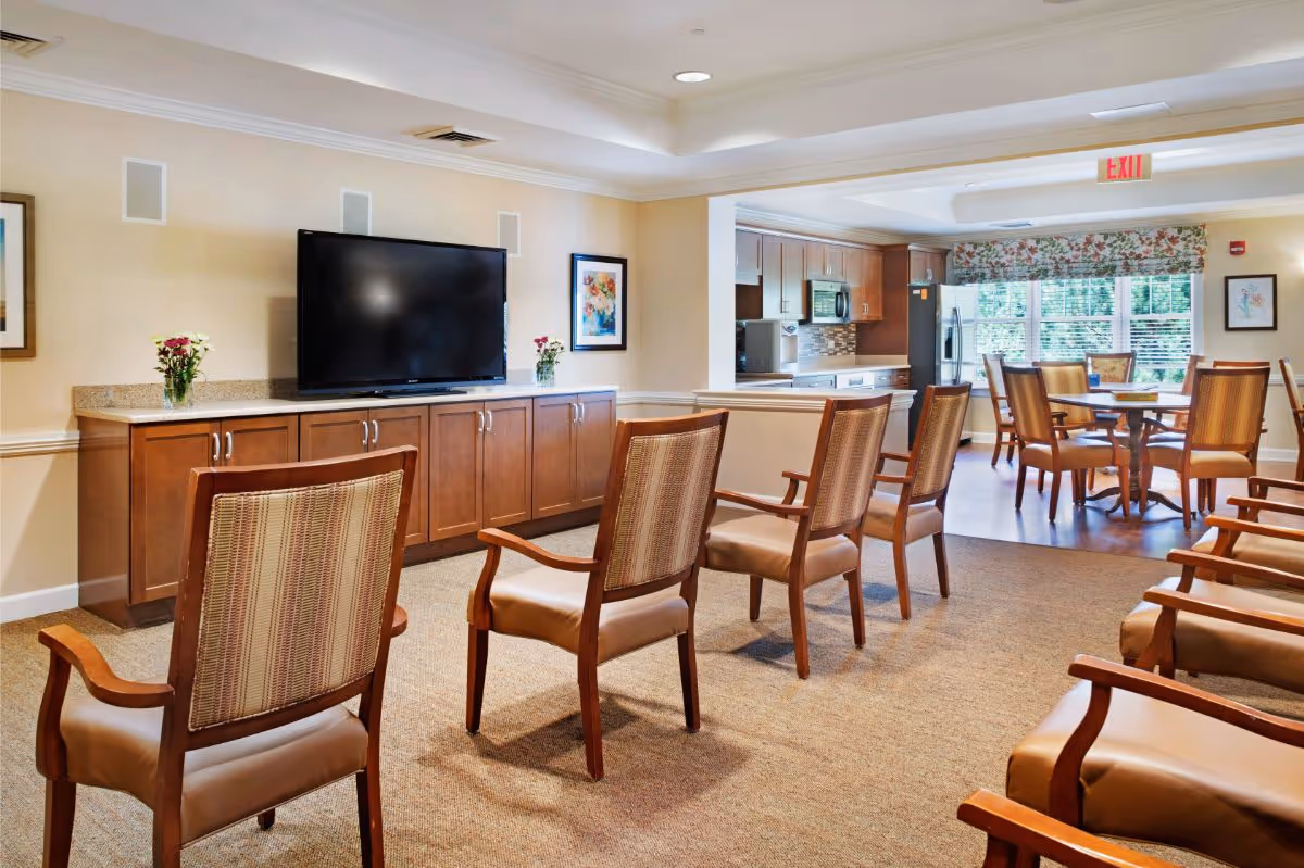 A bright communal sitting area with wooden chairs arranged facing a wall-mounted TV and a view into an adjacent kitchen and dining space.