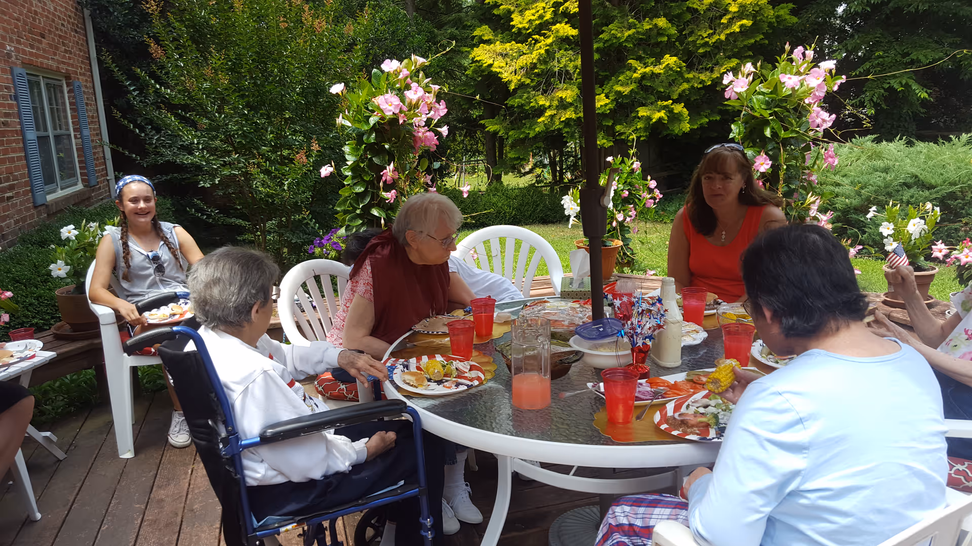 A group of people gathered around a round glass patio table eating a meal on a wooden deck surrounded by potted flowering plants.