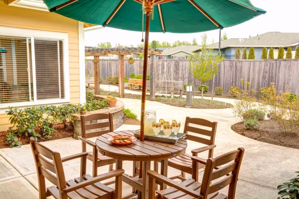 Outdoor patio area with a round wooden table and four wooden chairs under a large green umbrella. On the table, there is a pitcher of lemonade, four glasses with lemon slices, and a plate of cookies. The patio is surrounded by a garden with small plants and shrubs, a wooden fence, and a pergola in the background.