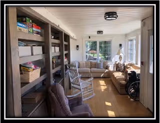 A bright living room with large windows letting in natural light. The room features a beige sectional sofa with cushions, a rocking chair, a wheelchair, and a bookshelf filled with board games and baskets. The floor is wooden, and the ceiling has two modern light fixtures.