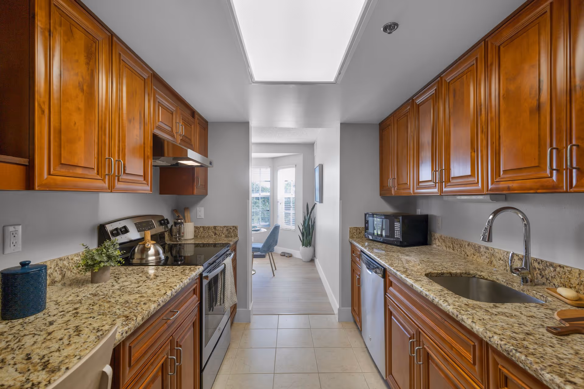 A galley kitchen with wooden cabinets and granite countertops on both sides. On the left side, there is a stove with a kettle and some kitchen utensils. On the right side, there is a sink, a microwave, and a dishwasher. The kitchen leads to a bright dining area with a window and a plant in the corner.