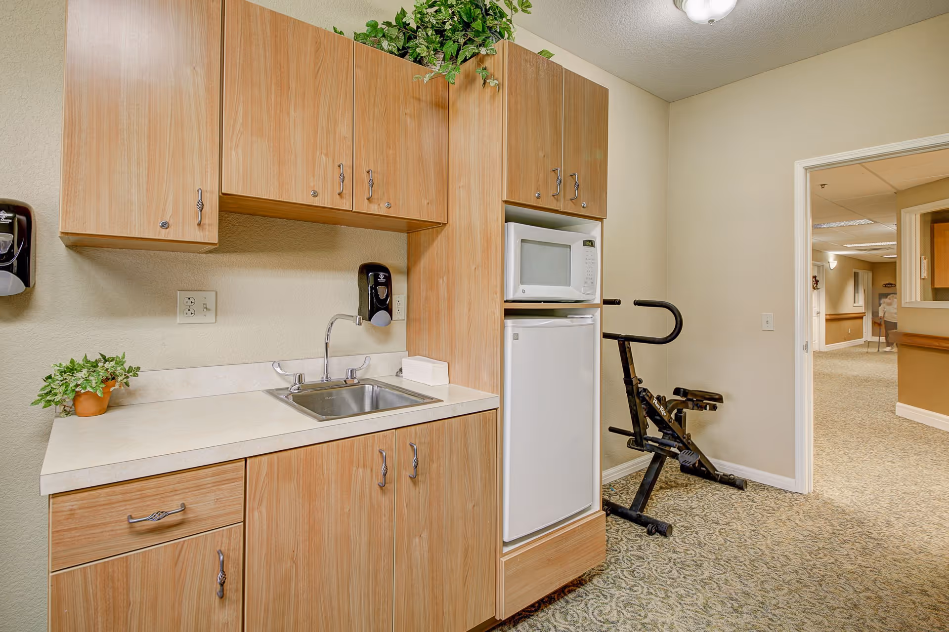 A small kitchenette area with light wood cabinets, a countertop with a sink, a microwave, and a mini refrigerator. There is a small potted plant on the counter and a wall-mounted soap dispenser above the sink. In the background, there is an exercise bike and a hallway with beige walls and patterned carpet.