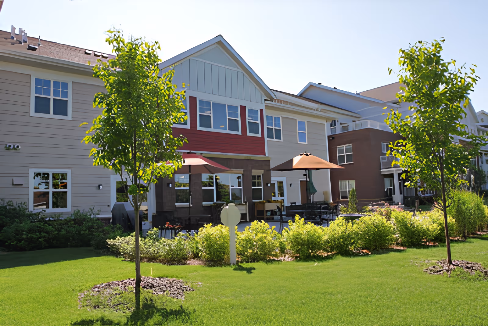 Exterior view of a Northern Lakes Senior Living building with a lawn, trees, and a patio area with umbrellas.