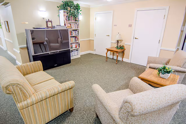 A cozy living room area with a large flat-screen TV on a stand, a bookshelf filled with books and decorative items, a small wooden side table with a lamp and plant, and three upholstered chairs arranged around a wooden coffee table with a plant on it. The walls are light-colored with white trim, and there are two closed white doors in the background.