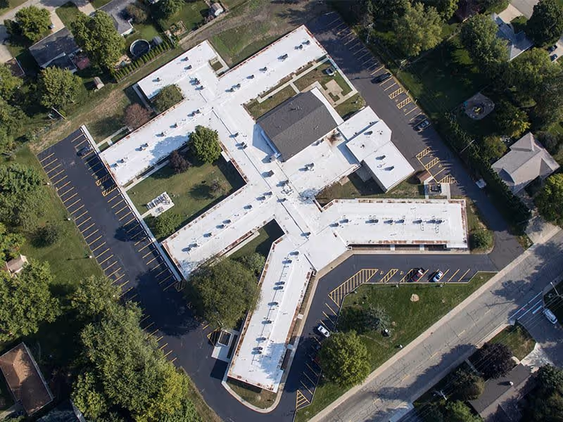 Aerial view of Morton Estates senior living facility showing a large, white-roofed building with multiple wings forming a Y shape, surrounded by parking lots, trees, and nearby residential houses.