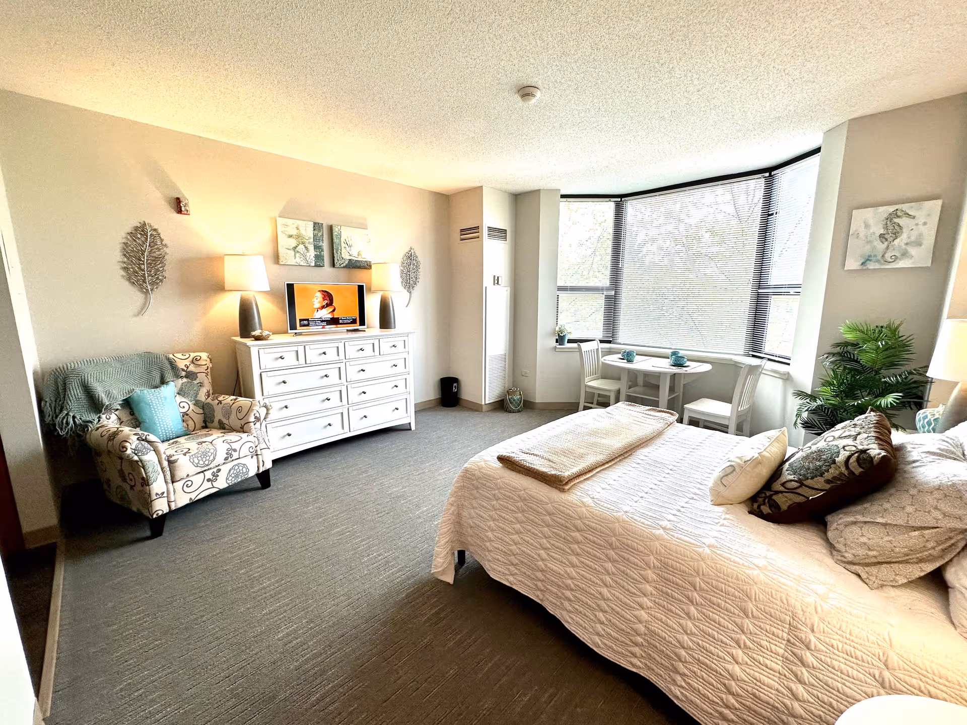 A bright and cozy bedroom in a retirement facility featuring a large bed with multiple pillows and a folded blanket, a patterned armchair with a throw blanket and cushion, a white dresser with a TV on top, two table lamps, wall decorations, a small round table with two chairs near a large window with blinds, and a potted plant in the corner.