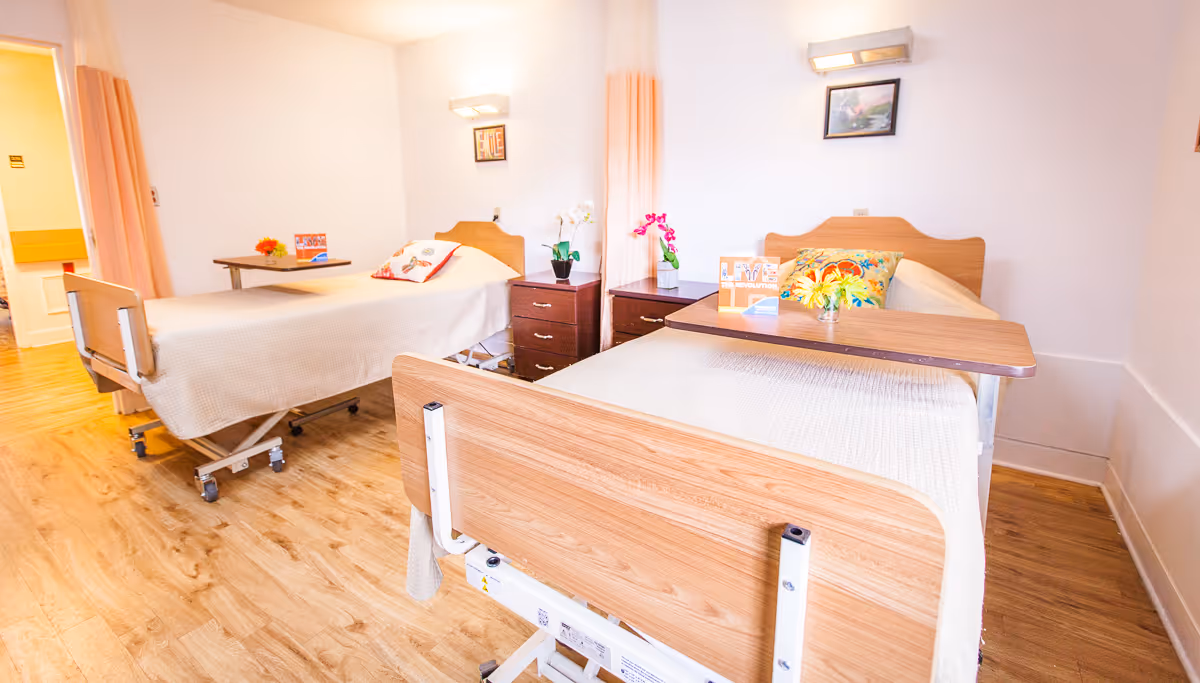 Bright shared patient room with two adjustable hospital beds, overbed tables, a nightstand, and floral decorations.