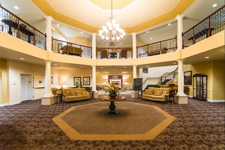 Spacious two-story senior living lobby with a central table of flowers, chandelier, seating areas, and upper balcony railings.