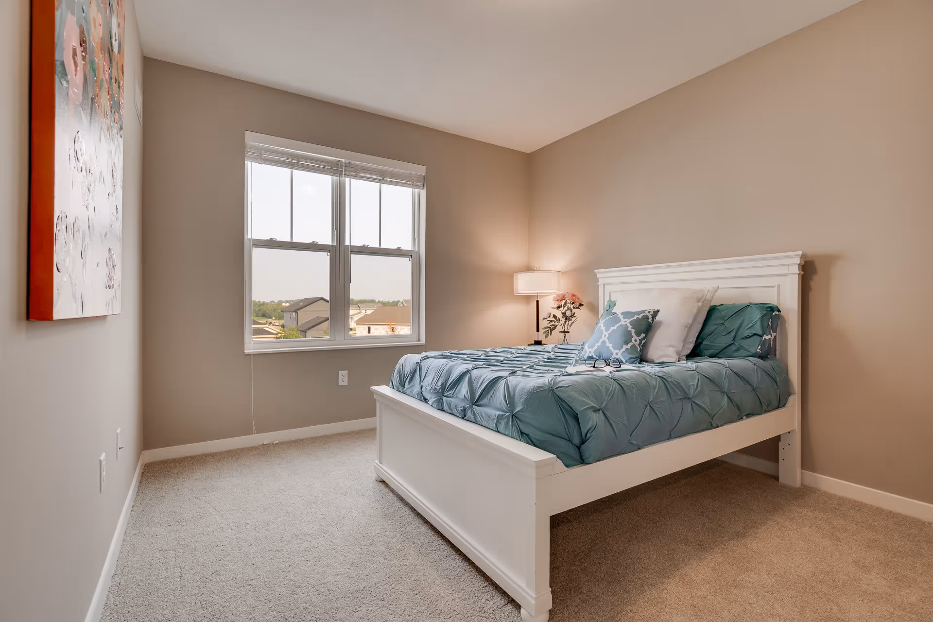 Sunlit bedroom with a white bed frame, teal bedding, a window, bedside lamp, and wall art.