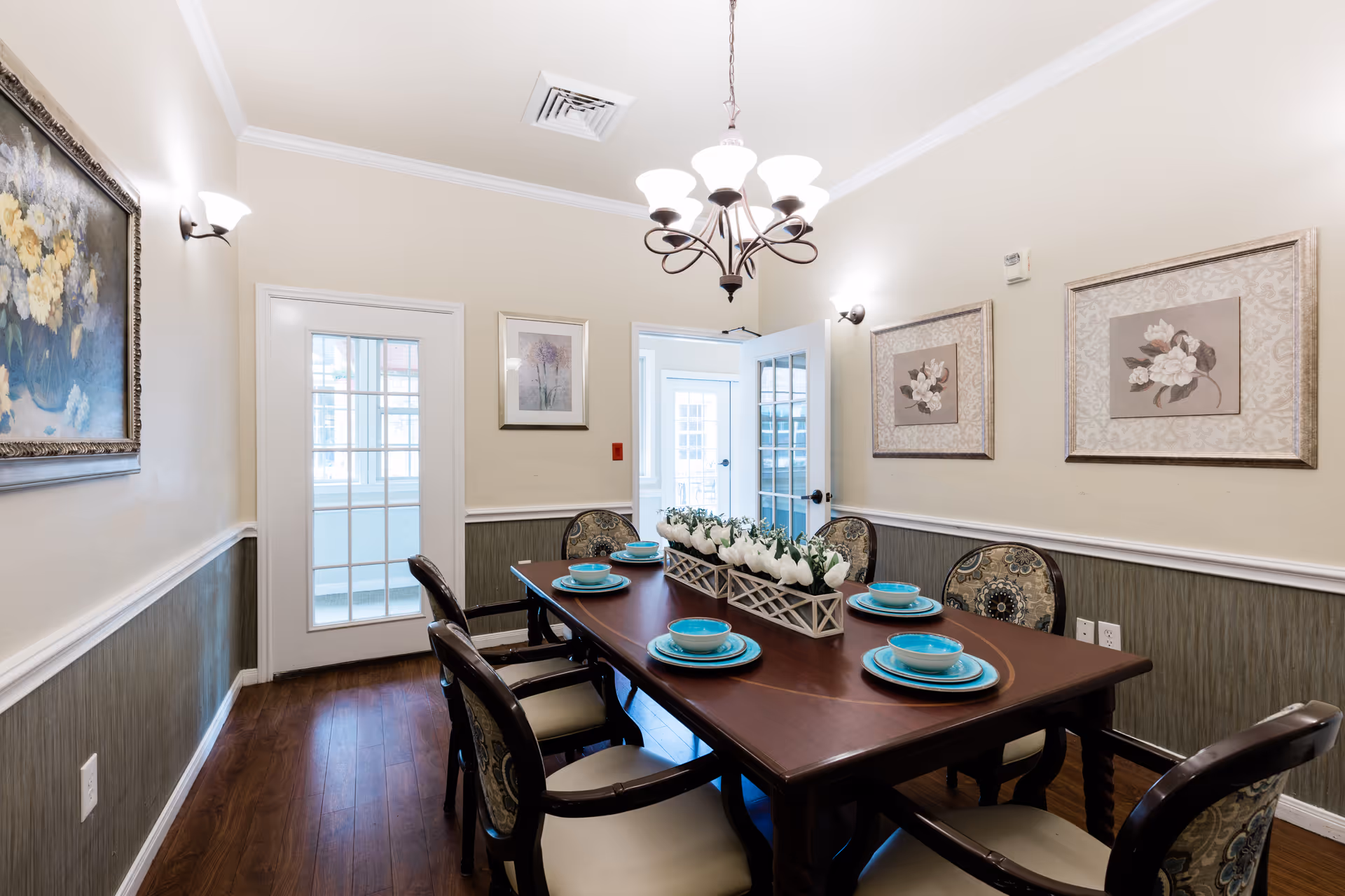 Formal dining room with a dark wood table set with turquoise dishes, a floral centerpiece, upholstered chairs, framed artwork and a chandelier.