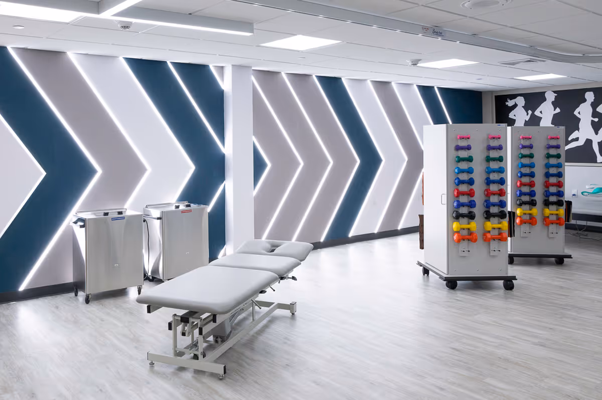 A modern rehabilitation room with a treatment table in the center, two stainless steel carts, and two racks holding colorful dumbbells. The back wall features a striking design of illuminated chevron patterns and a black and white silhouette mural of running figures.