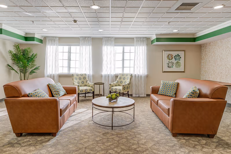 A bright and spacious living room area with two brown leather sofas facing each other, each with patterned throw pillows. Between them is a round coffee table with a small plant centerpiece. In the background, there are two patterned armchairs with a small round table between them, positioned in front of three large windows with sheer white curtains. A green potted plant is on the left side, and a framed botanical artwork hangs on the right wall. The room has a beige carpet and a white ceiling with recessed lighting.