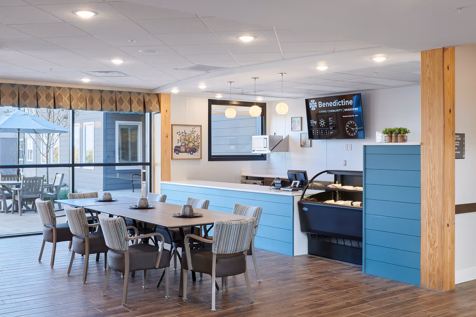 A bright dining area in a senior living community featuring a long wooden table with eight cushioned chairs. The room has large windows with a view of an outdoor patio with tables and umbrellas. The interior includes a blue and white counter area with a display case, a mounted TV screen showing the Benedictine Living Community logo and information, and pendant lights hanging from the ceiling.