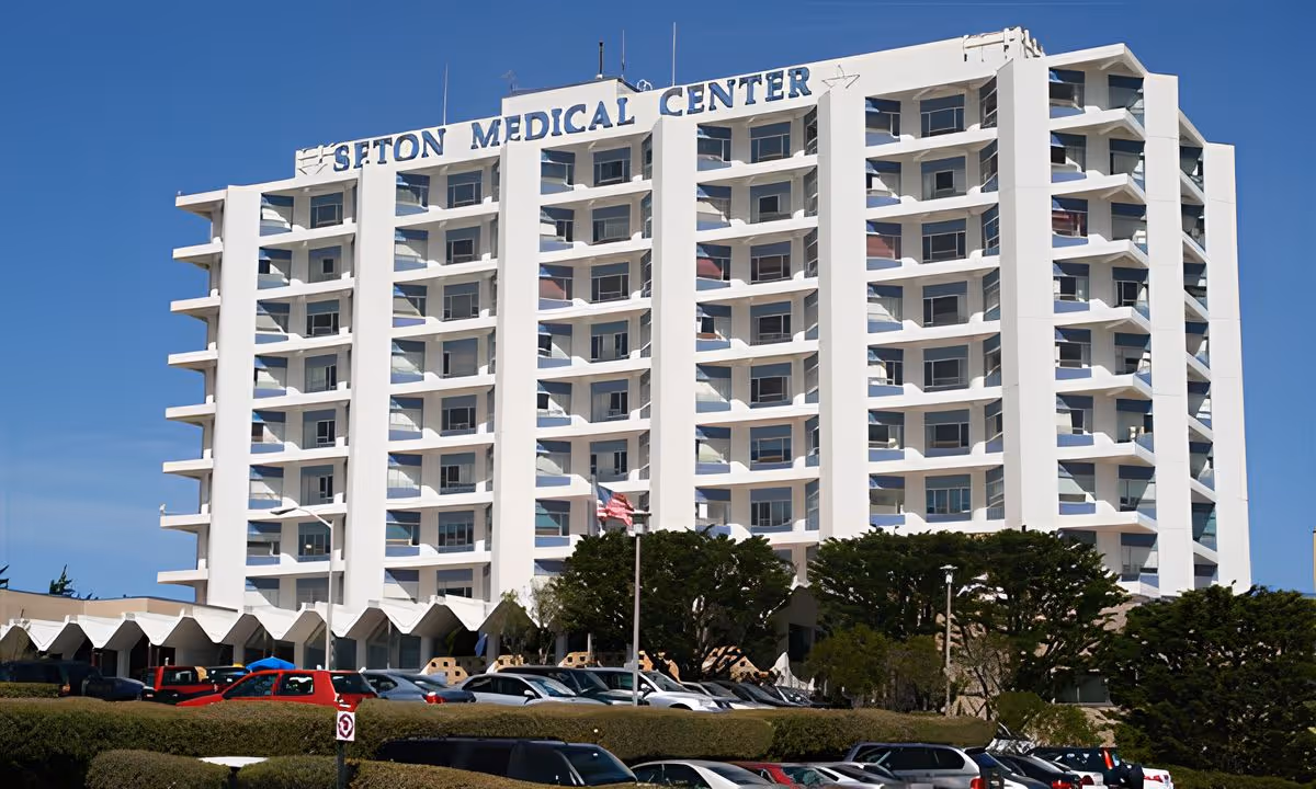 Front view of the multi-story white Seton Medical Center building with balconies, a parking lot and trees in front under a clear blue sky.