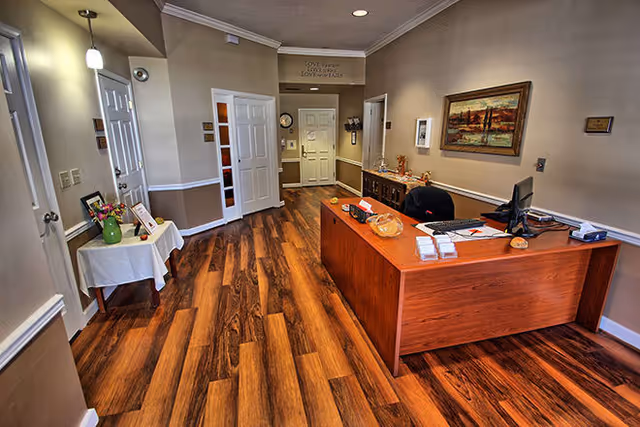 Reception area with a wooden desk, computer, and office chair. The room has wood flooring, beige walls with white trim, a small table with flowers and framed photos, and a painting on the wall. Multiple doors and a clock are visible in the background.