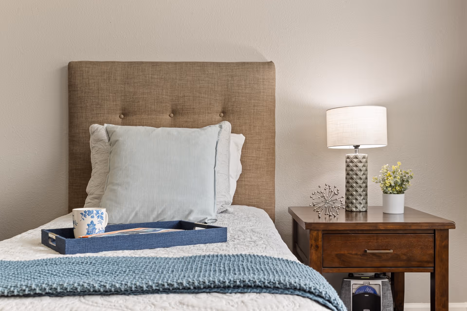 A neatly made bed with a brown upholstered headboard, white and light blue pillows, and a blue knitted throw blanket at the foot. A blue tray with a white and blue floral mug and a magazine rests on the bed. Next to the bed is a wooden nightstand with a decorative lamp, a small metal sculpture, and a small potted plant with yellow flowers.