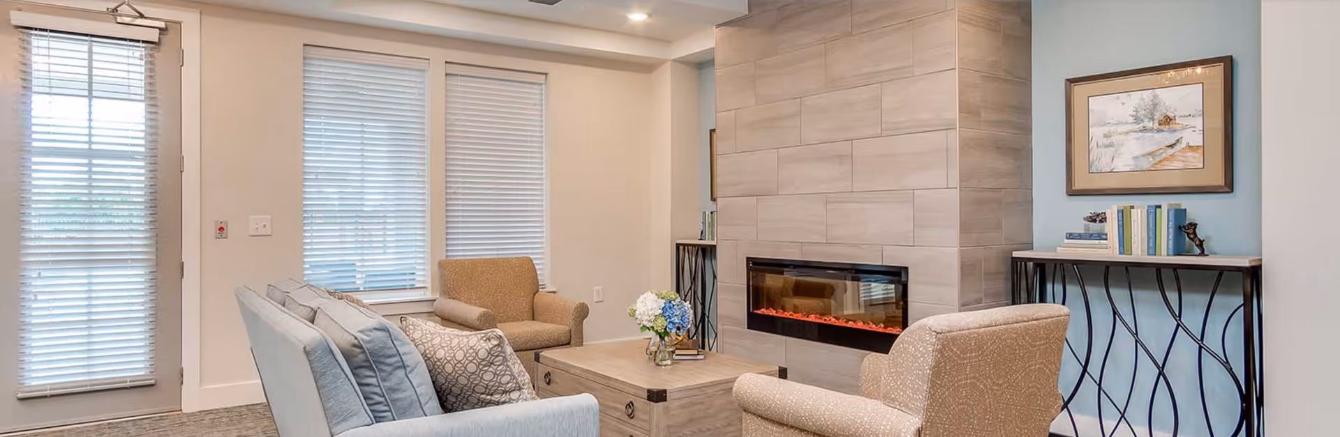 Cozy living room with sofas and armchairs arranged around a modern wall-mounted fireplace and a coffee table near windows with blinds.