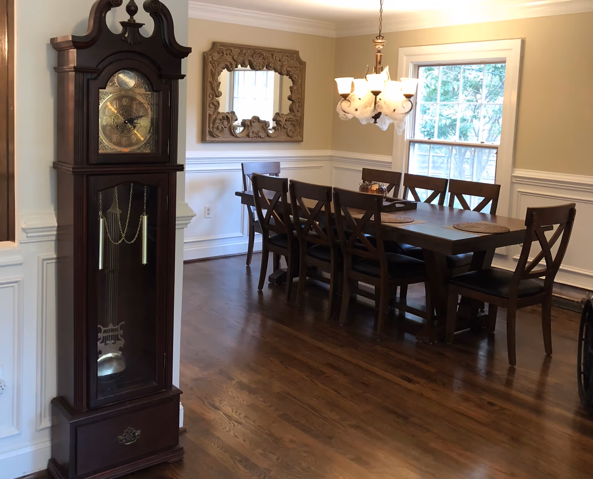 A dining room with a long wooden table surrounded by eight matching wooden chairs with black cushions. A decorative chandelier hangs above the table. On the left side, there is a tall grandfather clock with a glass front and ornate clock face. A large ornate mirror is mounted on the wall behind the table. The room has wooden flooring, beige walls with white wainscoting, and a window letting in natural light.