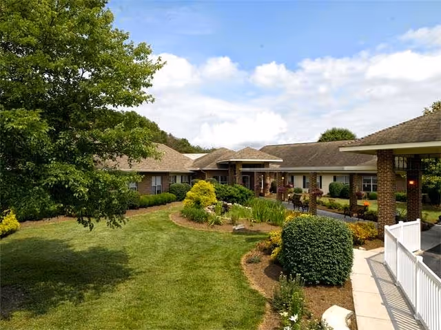 View of a senior living facility building with a well-maintained lawn, landscaped garden beds, bushes, and trees under a partly cloudy sky.