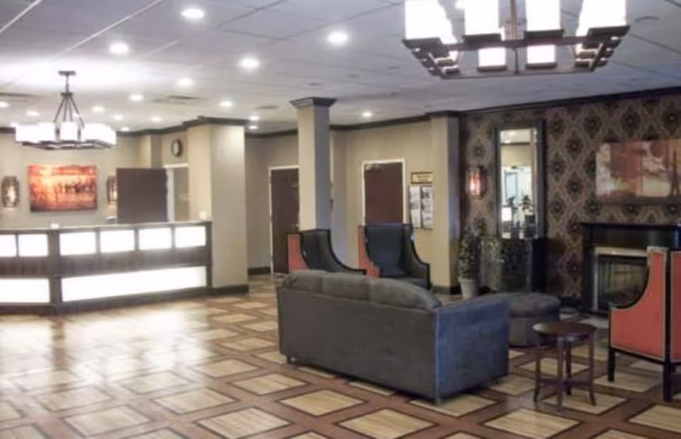 A spacious and well-lit lobby area with a reception desk on the left, a seating area with a gray sofa and two red and black armchairs around a small table, decorative wallpaper with a mirror and artwork above a fireplace, and a patterned tile floor.
