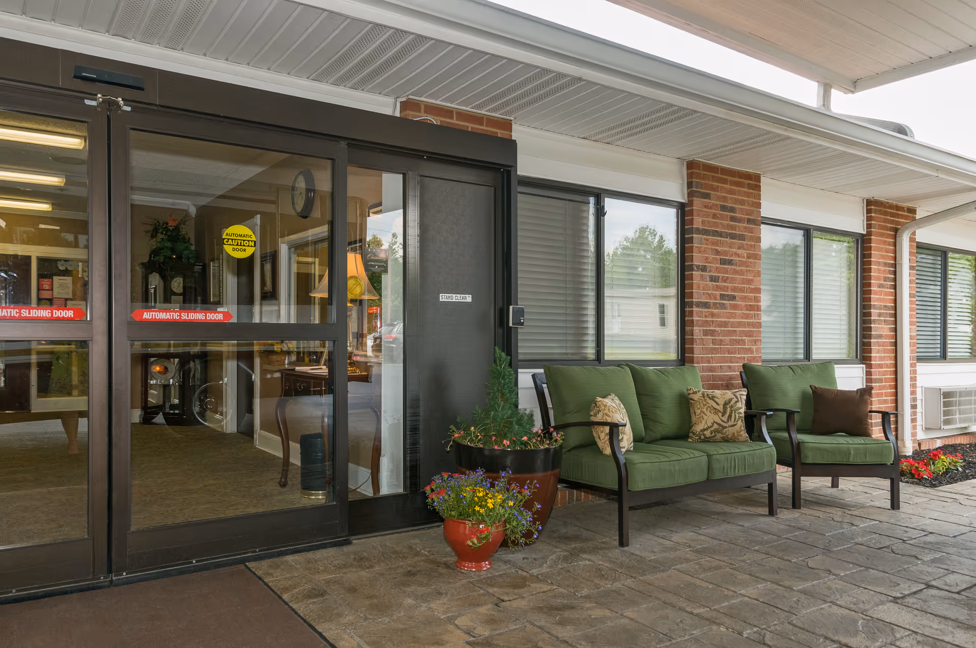 Entrance area of a senior living facility with automatic sliding glass doors. Outside the entrance, there are two green cushioned chairs and a matching loveseat with decorative pillows. There are also two flower pots with colorful flowers and a brick wall with windows in the background.