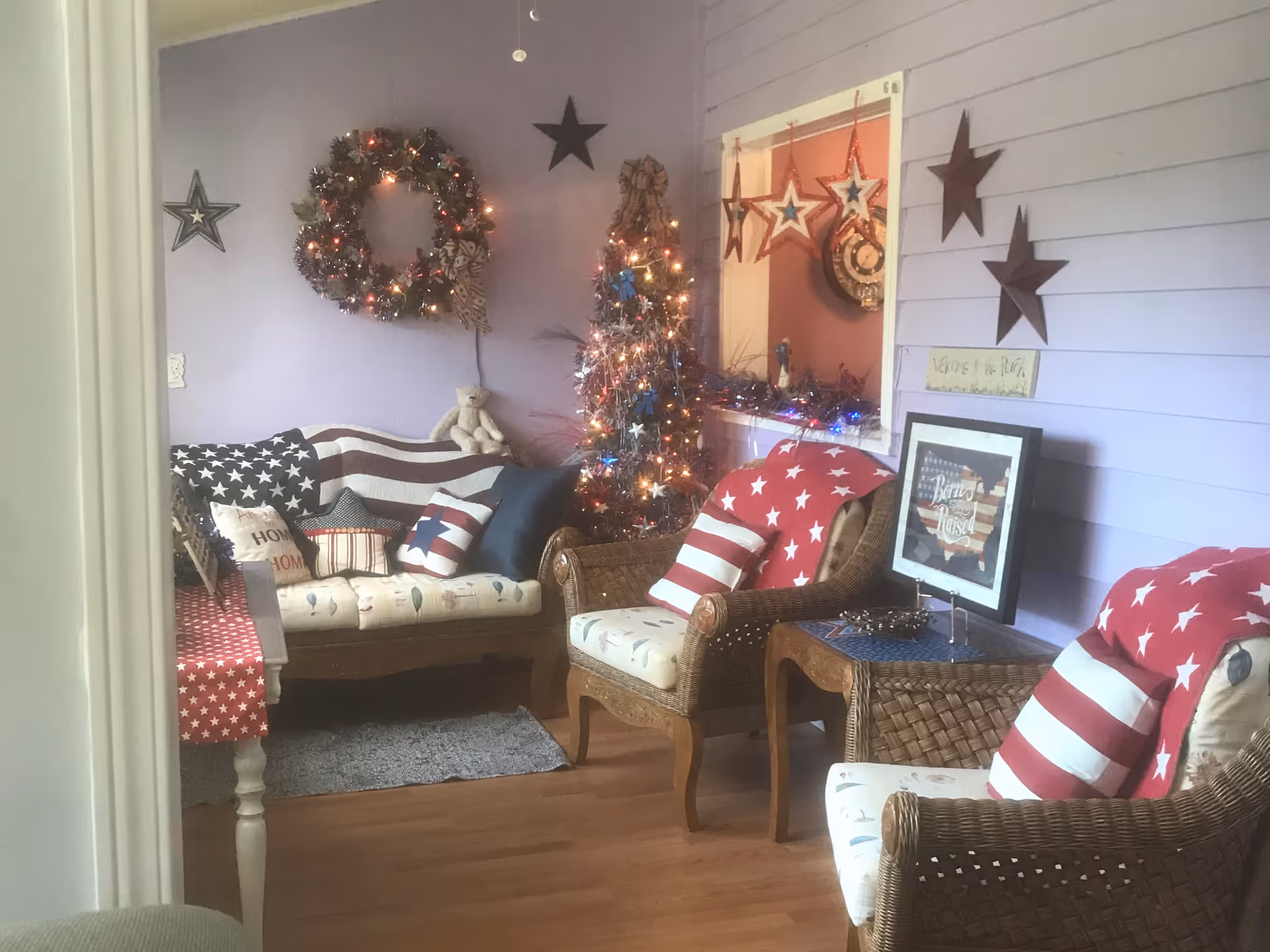 A cozy living room decorated with patriotic American flag-themed pillows and blankets. The walls are painted light purple and adorned with star decorations and a lit wreath. There is a small decorated Christmas tree with lights, a wicker armchair, a wicker side table with a framed picture, and a cushioned sofa with various pillows. The room has wooden flooring and a small gray rug.