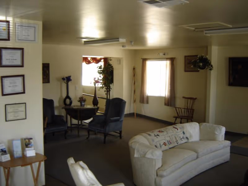 A cozy living room area with a white couch adorned with a patterned pillow, two dark armchairs around a small round table with decorative vases, a wooden chair near a window with curtains, framed certificates on the wall, and soft lighting from ceiling fixtures.