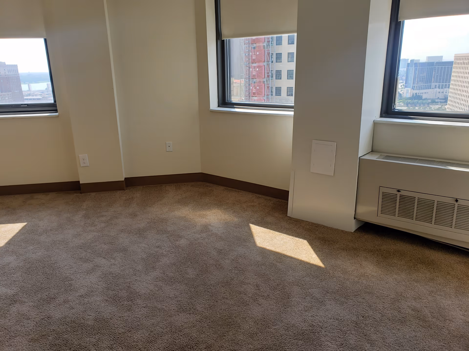 Empty corner of a room with beige carpet, cream-colored walls, two windows with city views, and a heating/cooling unit below one window.