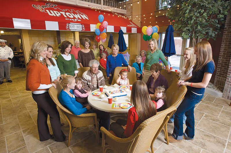 A group of people of various ages, including seniors, adults, and children, gathered around a round table with a birthday cake and colorful balloons in a festive indoor setting resembling a cafe or dining area with a red awning labeled 'Uptown Cafe'.