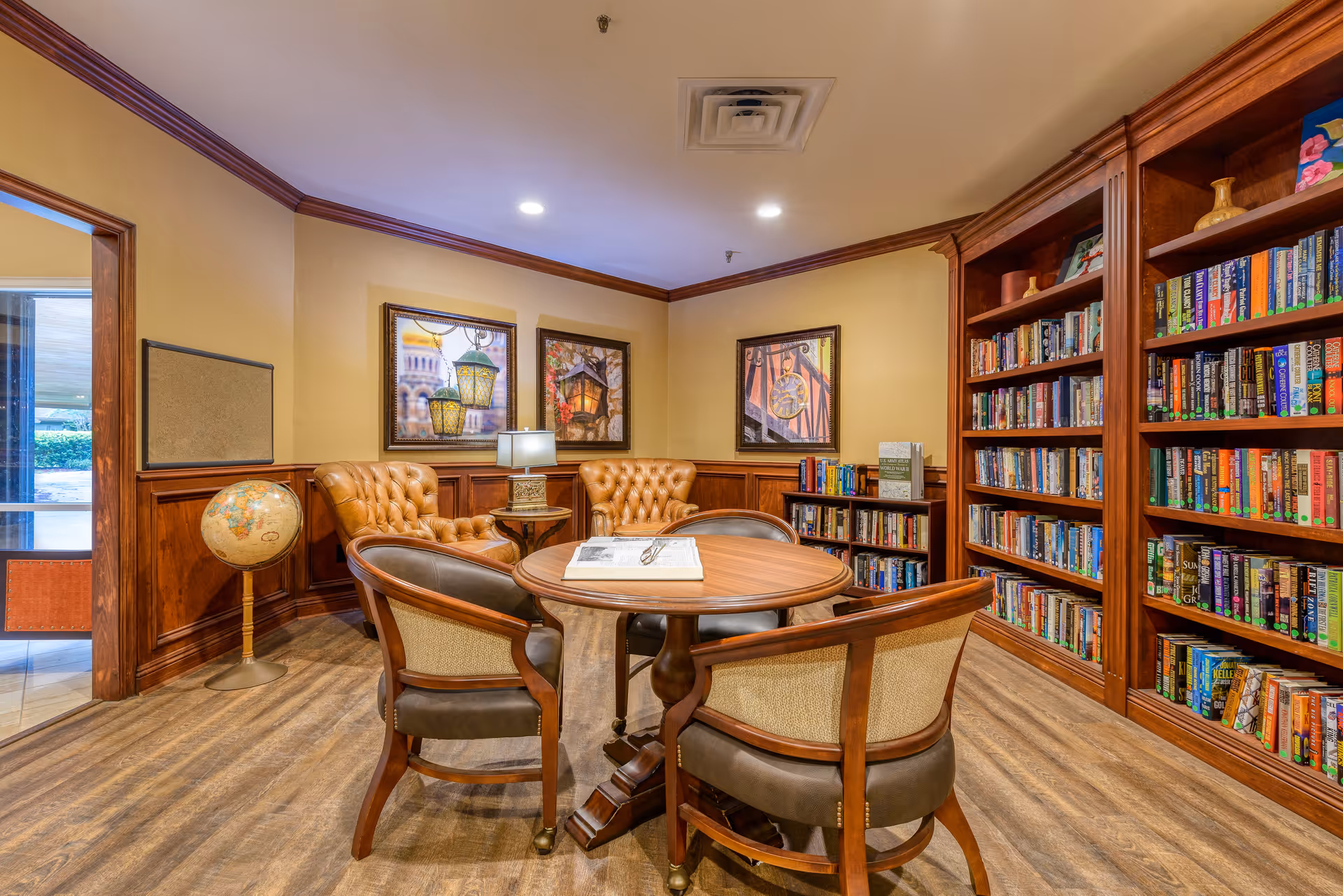 A cozy library room with wooden bookshelves filled with books, a round wooden table with four chairs, two tufted leather armchairs, a small side table with a lamp, a globe on a stand, and framed artwork on the walls.