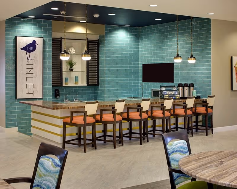 Interior view of a modern dining area with a long counter featuring eight high chairs with orange cushions. The back wall is covered with teal subway tiles and has a mounted TV, coffee dispensers, and a small shelf with decorative items. Pendant lights hang above the counter, and there is a sign on the wall that reads 'THE INLET'. In the foreground, part of a round table and chairs with patterned upholstery are visible.