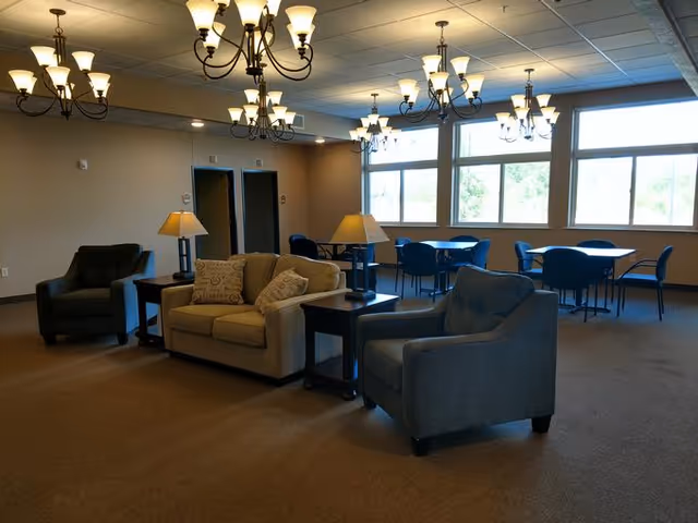 A spacious common area in an assisted living facility featuring a beige sofa with decorative pillows, two armchairs, side tables with lamps, and multiple chandeliers hanging from the ceiling. In the background, there are several tables with blue chairs near large windows letting in natural light.