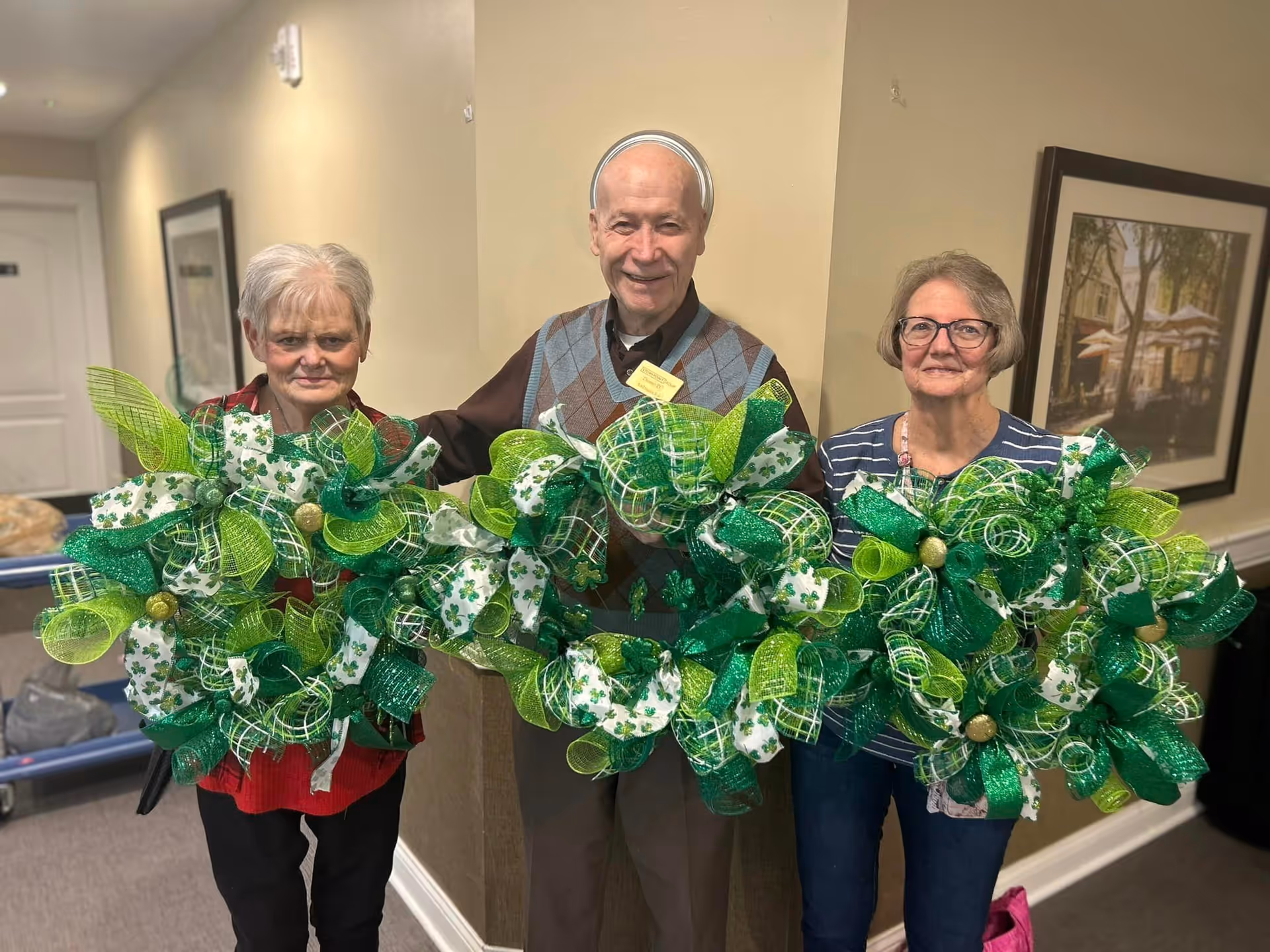 Three elderly individuals standing indoors holding large decorative wreaths adorned with green and white ribbons, some featuring shamrock patterns. They are smiling and appear to be in a hallway with framed artwork on the walls.